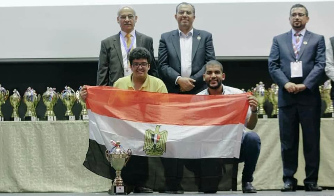 Holding the Egyptian flag with a teammate at a robotics awards ceremony, with a trophy in front