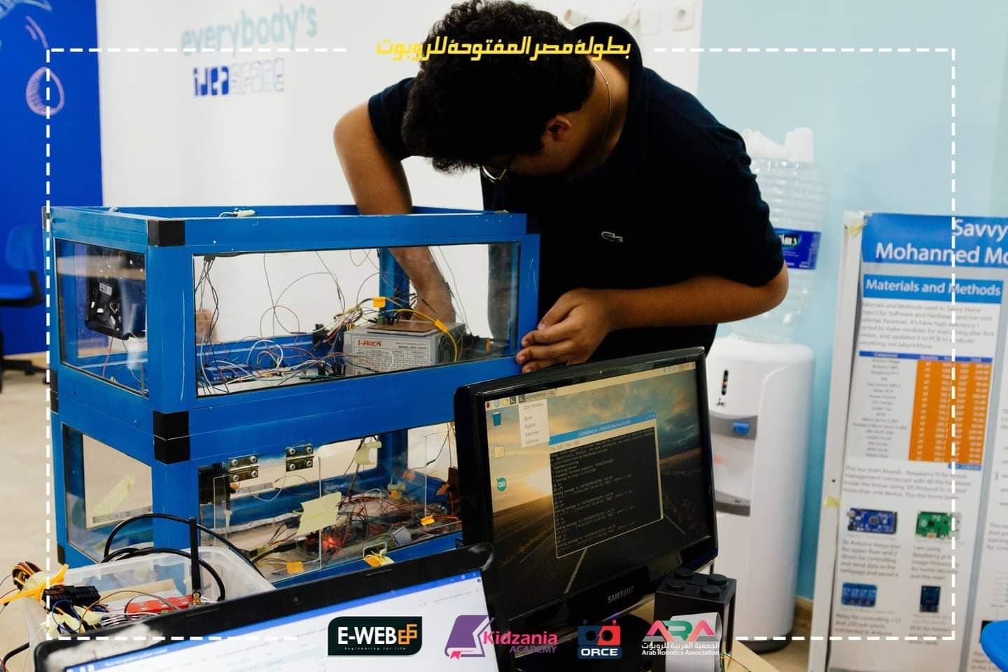 Leaning over my blue-framed smart-home prototype, with exposed wiring and computers beside it